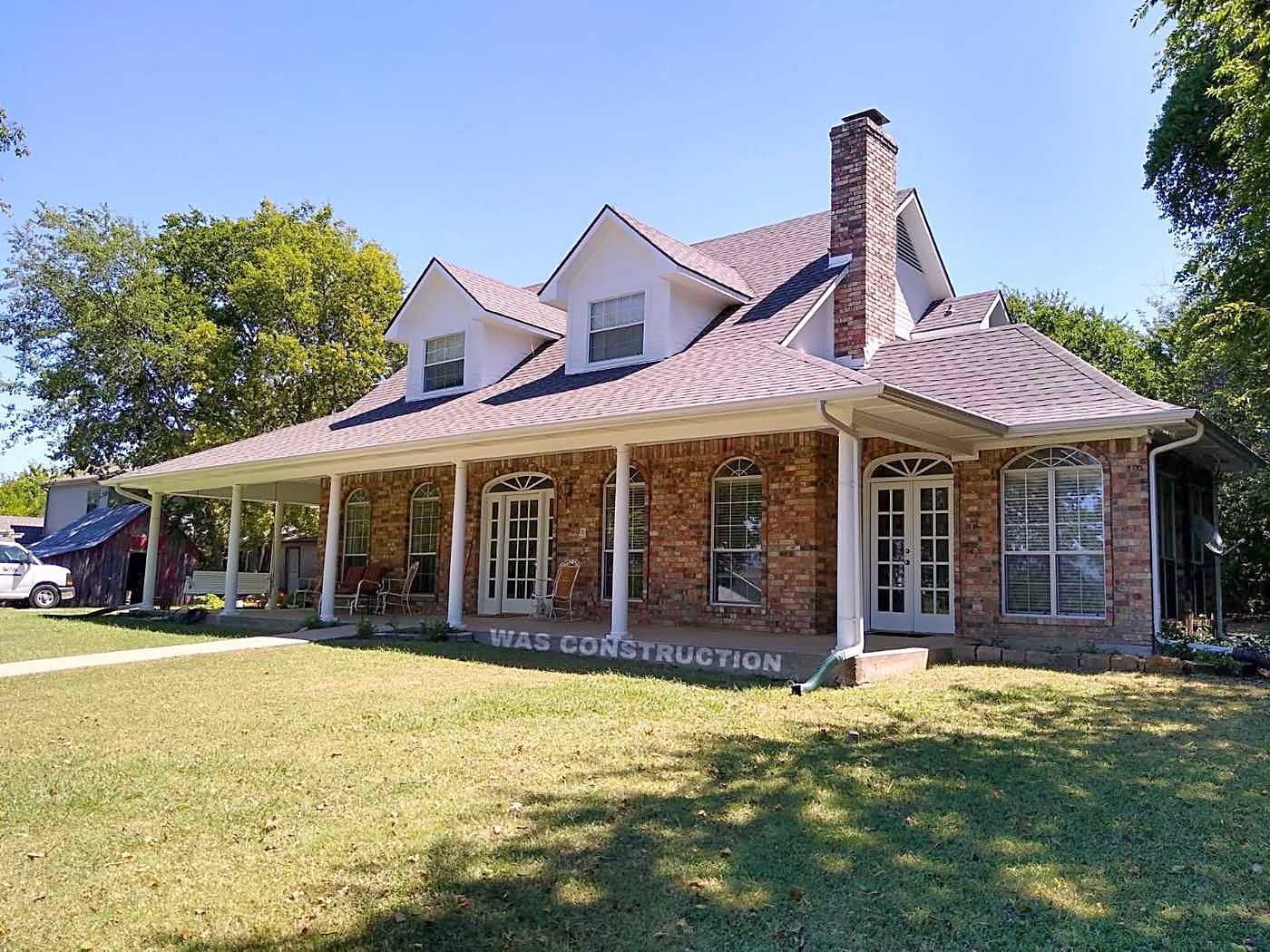 A brick home with a wrap-around porch and a finished shingle roof